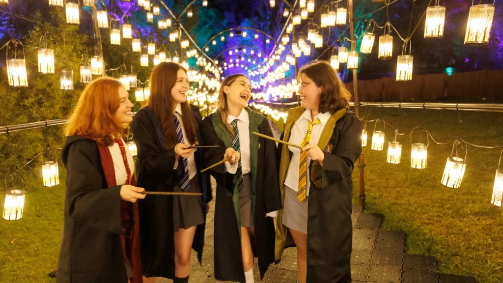 Four visitors in Hogwarts robes holding wands beneath rows of glowing lanterns at Harry Potter: A Forbidden Forest Experience